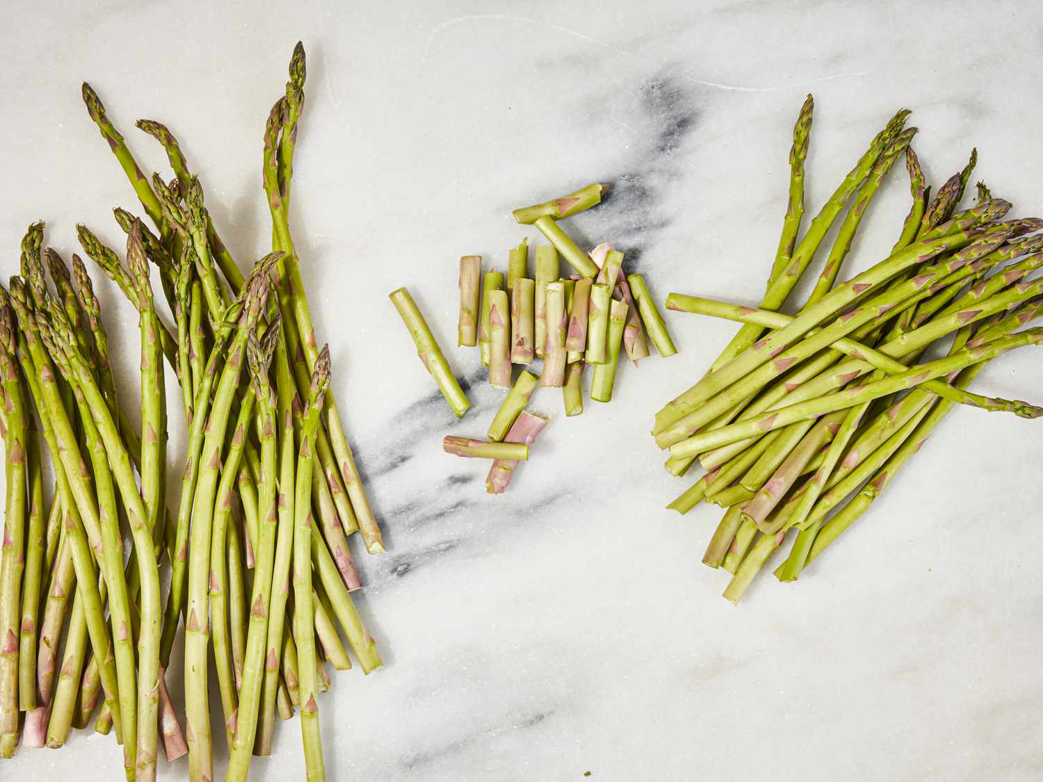 A pile of trimmed and untrimmed asparagus spears. The woody stem-end trimmings are in a small pile between them.