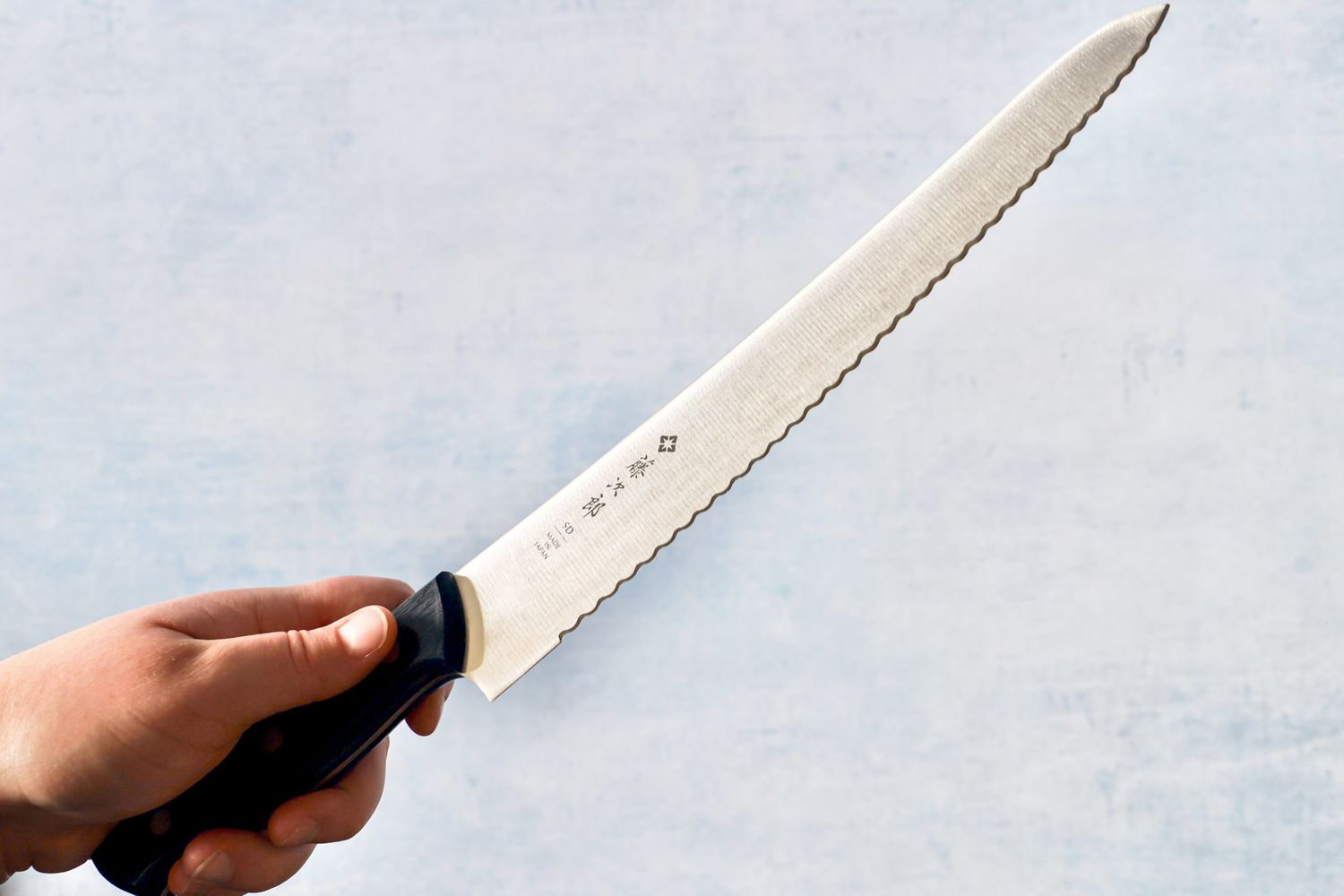 A hand holding the Tojiro F-687 Hand Made Bread Knife above a blue countertop background