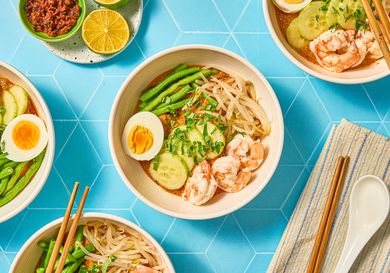 Bowls of curry laksa garnished with shrimp boiled egg cucumber and beans accompanied by lime and chili paste on a blue background