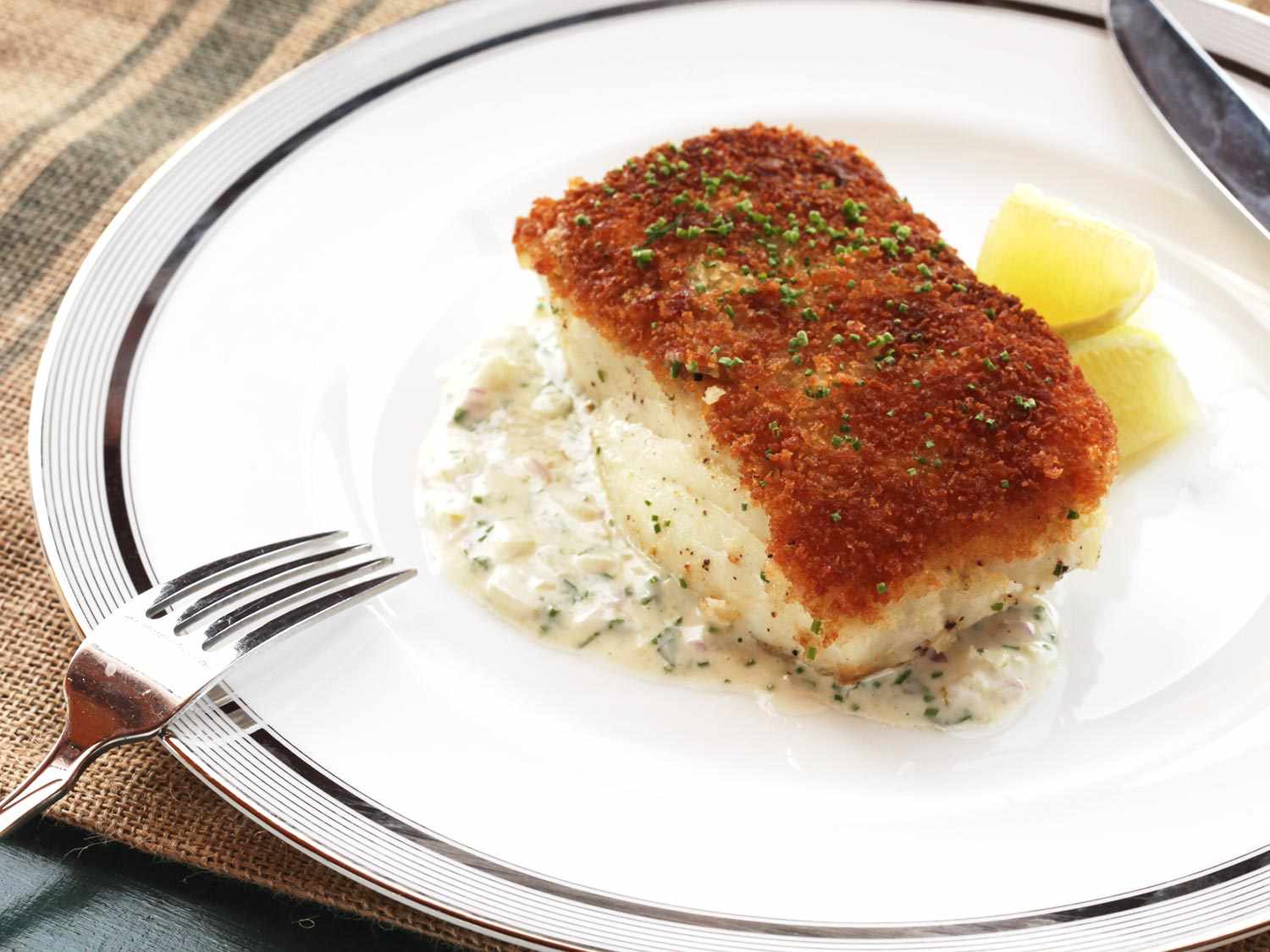 A plated fish fillet portion, panko crust facing up. The fish is sprinkled with minced chive, accompanied by lemon wedges, and positioned on a pool of tartar sauce.