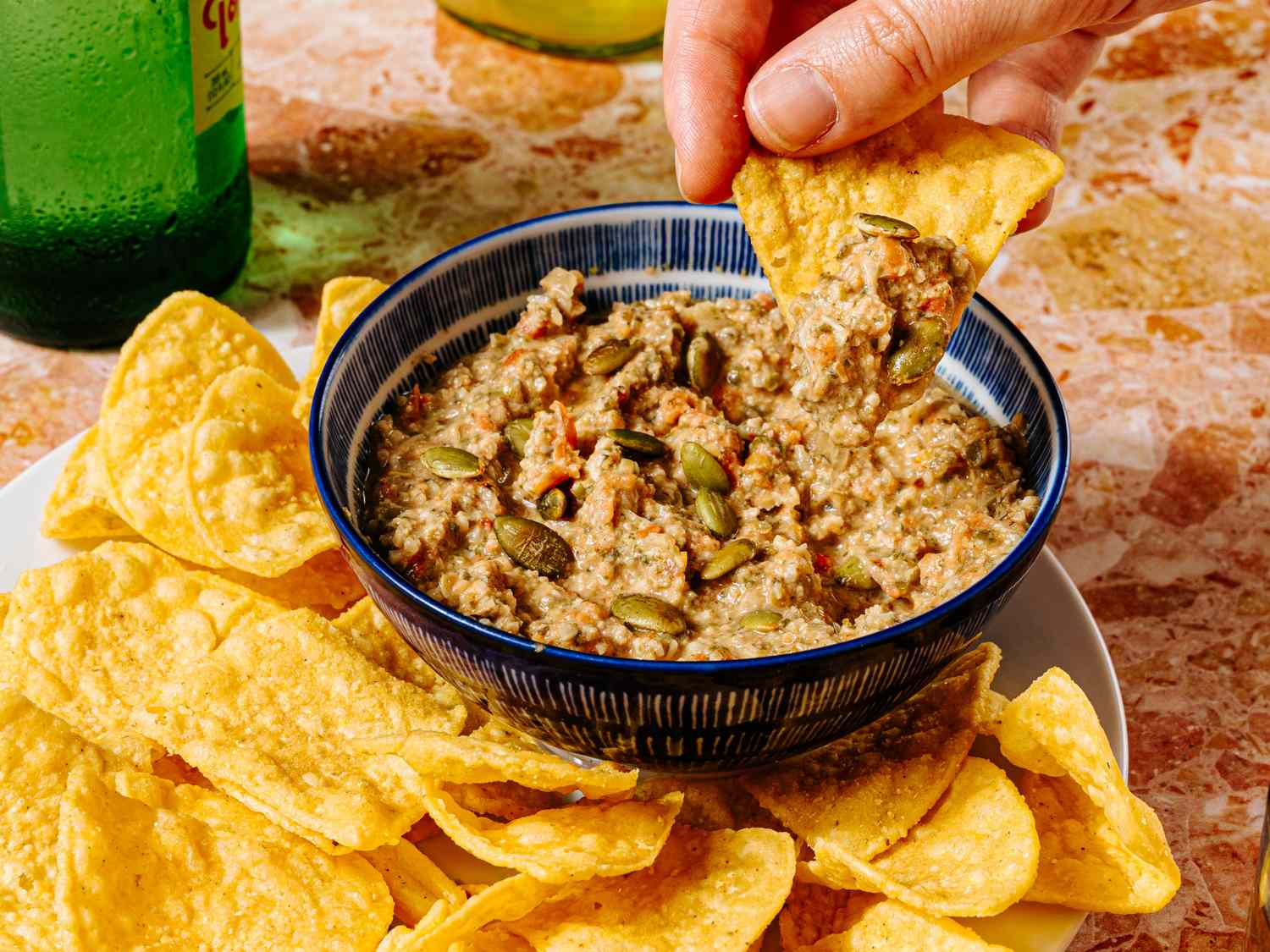 A hand dipping a chip into a bowl of dip accompanied by a plate of tortilla chips on a table