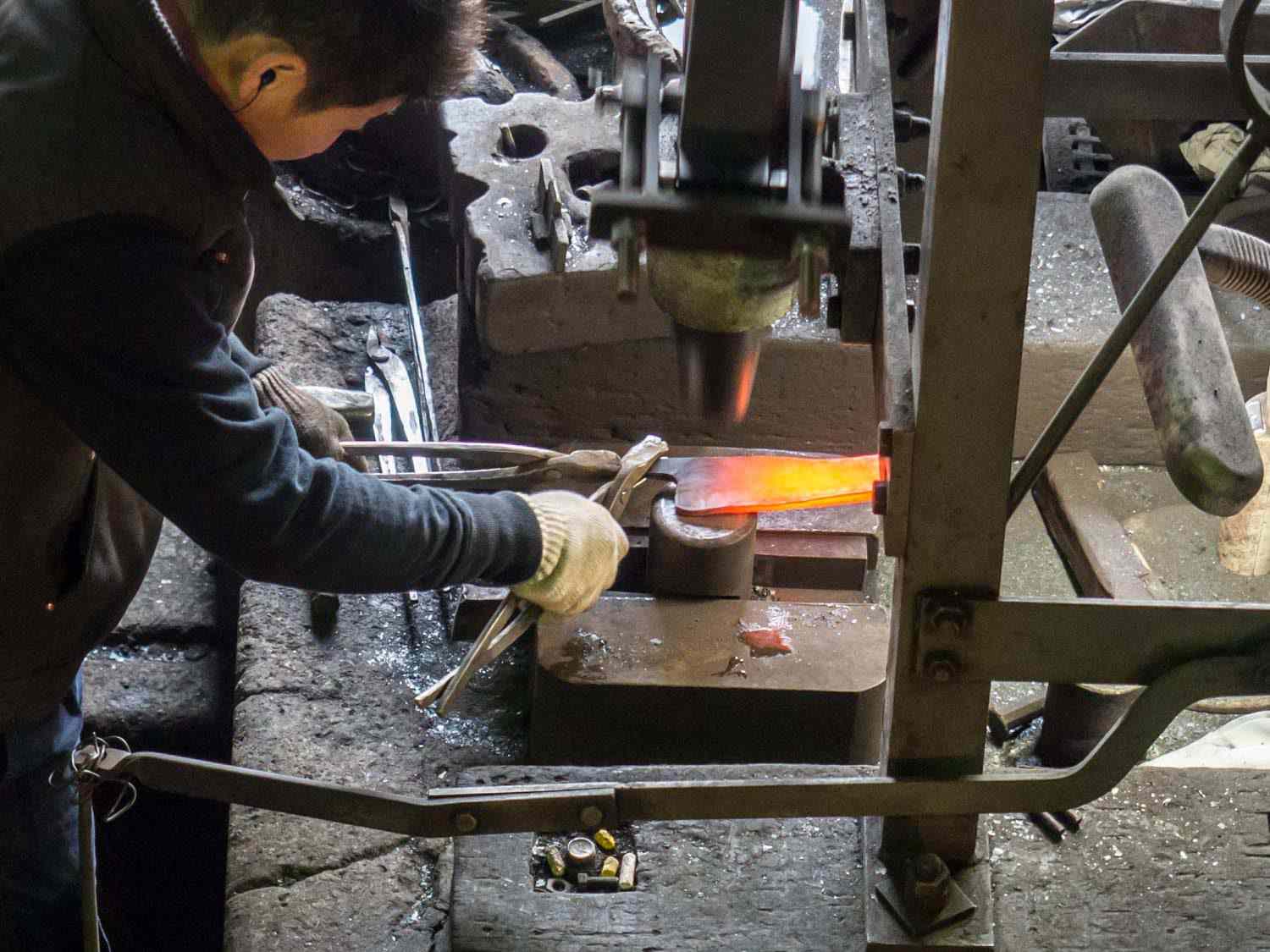 A Japanese bladesmith works with stacked, glowing knife blanks, hammering them so that they become thinner and uniform.