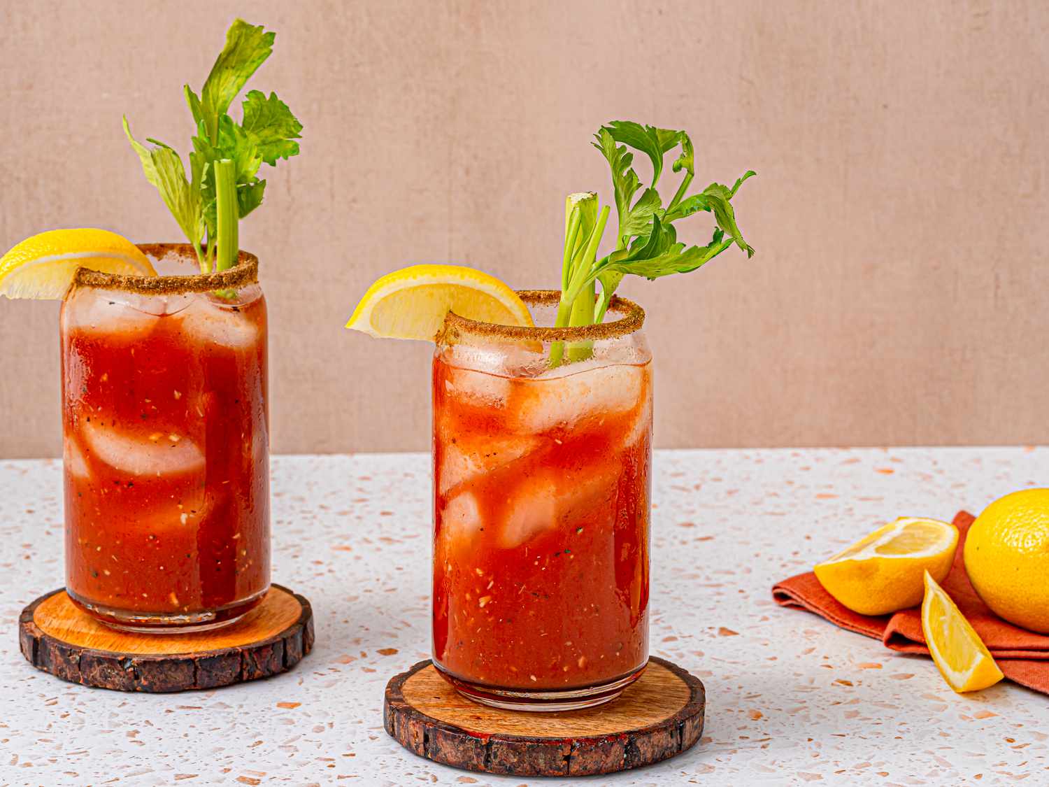 Two bloody Marys on wooden coasters garnished with celery and lemon wedges.
