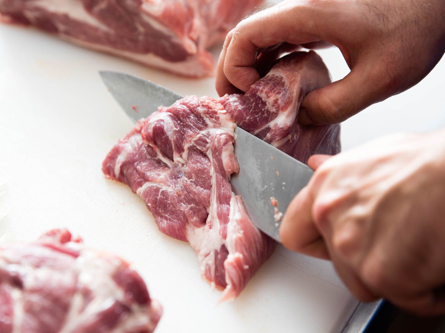 A large chunk of boneless pork shoulder is sliced thinly on a cutting board with a chef's knife.