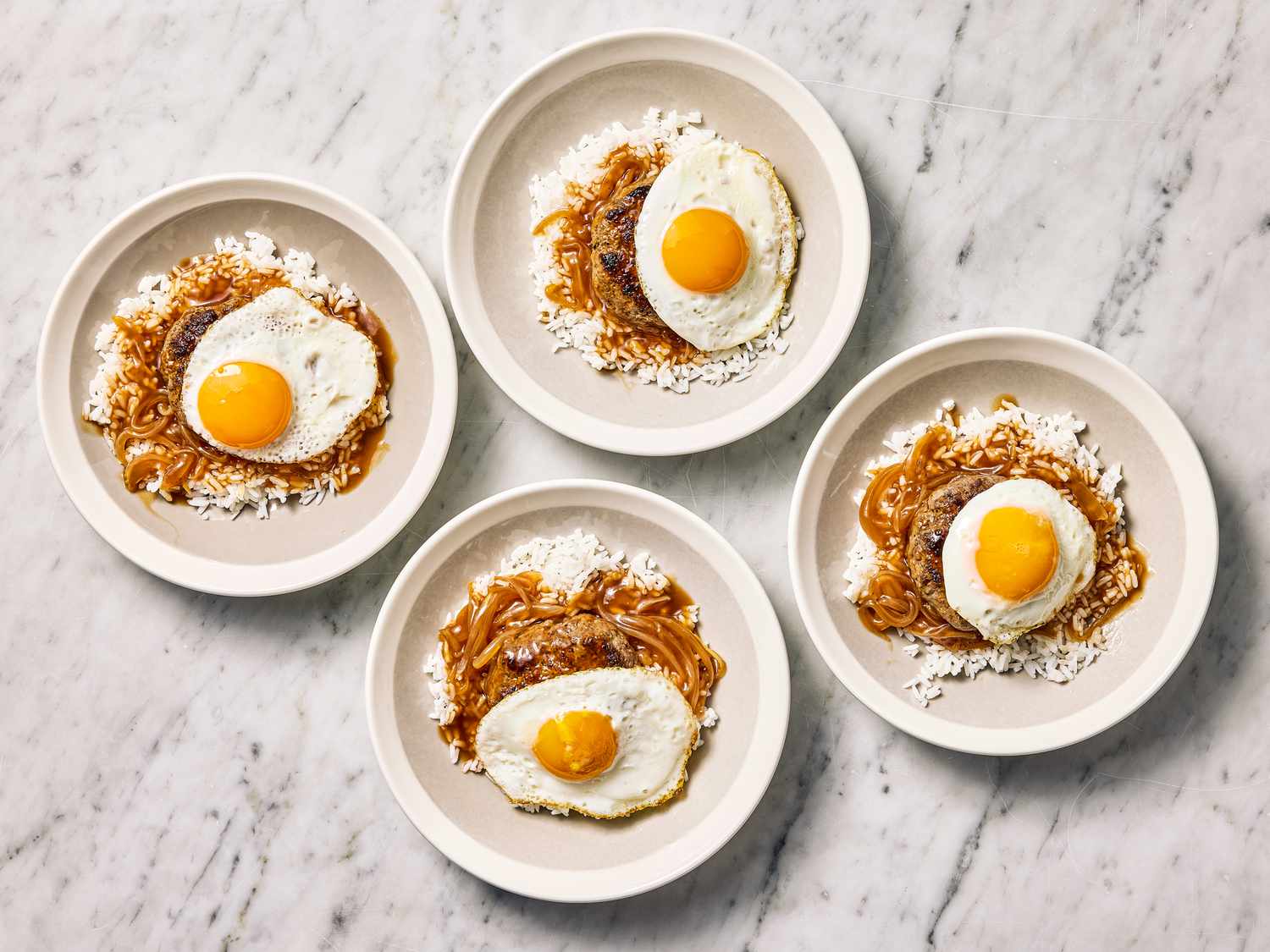 Four plates of food each with a fried egg on top, served on a marble countertop