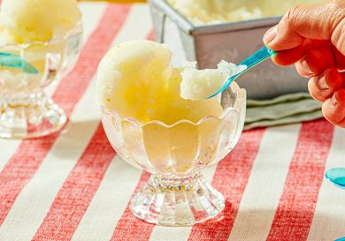 A hand scooping lemon sorbet from a glass bowl with a blue spoon placed on a red and white striped tablecloth