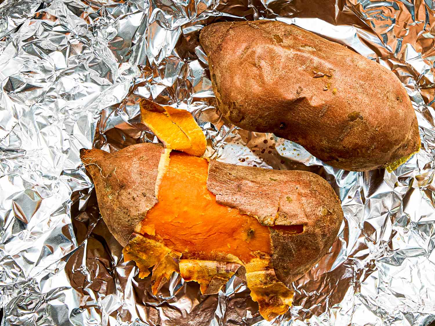 Two roasted sweet potatoes on foil one partially peeled to expose the orange flesh inside