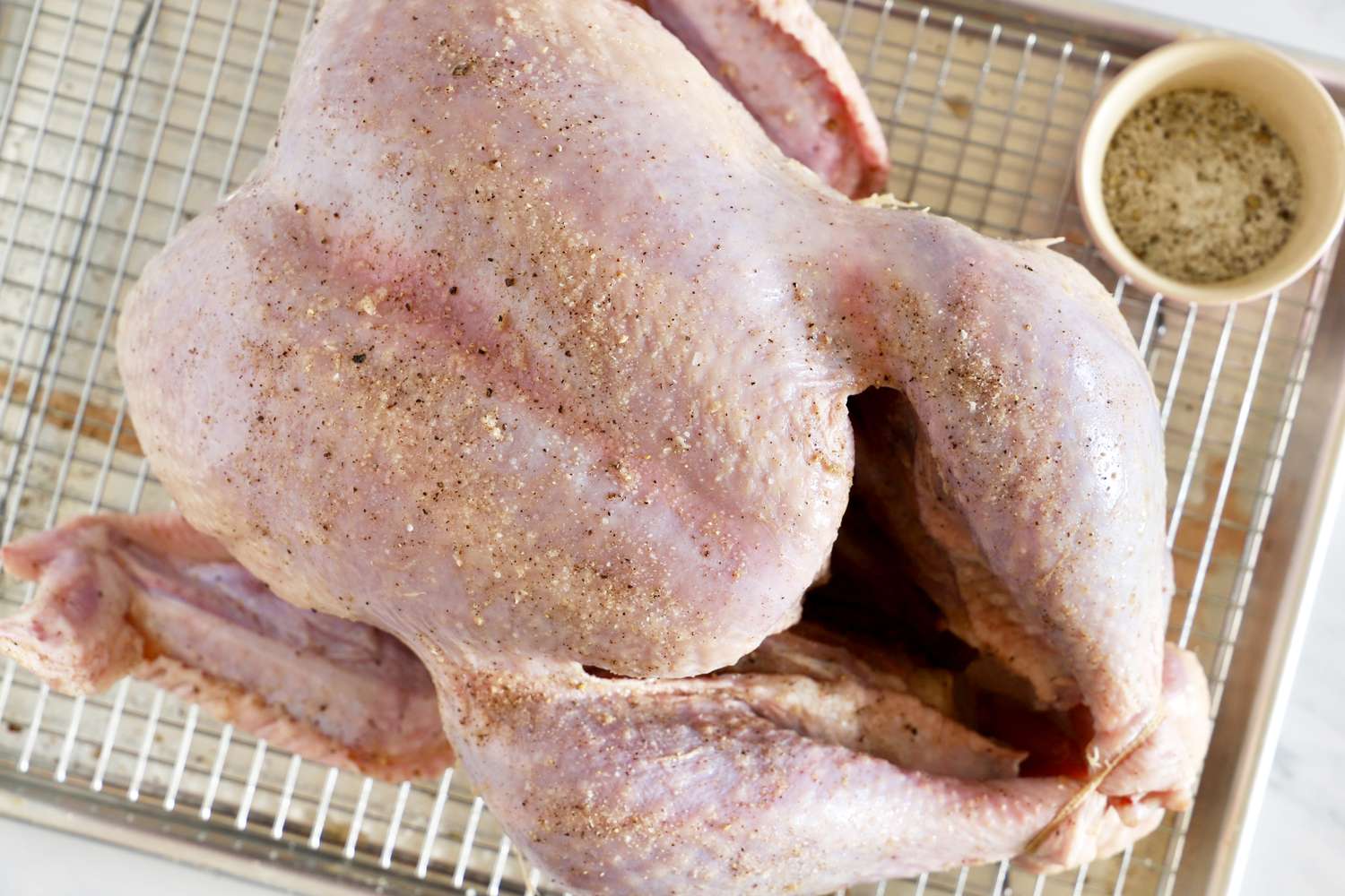 A raw seasoned turkey on a wire rack with a small cup of seasoning