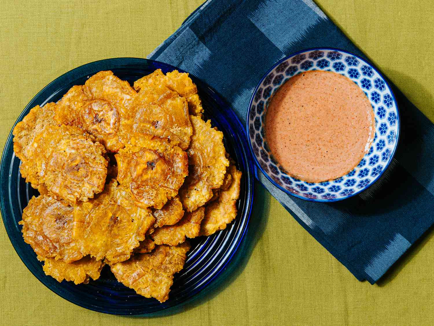 Overhead view of tostones on a blue plate with a blue napkin and bowl of dipping sauce
