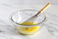 A whisk in a glass Pyrex bowl with a yellow liquid on a marble surface