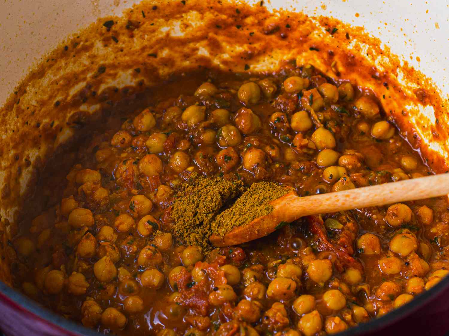 A close-up of the interior of the Dutch oven holding the channa masala. There is a wooden spoon with a portion of garam masala spice on top of it, showing that the spice is about to be stirred into the finished dish.