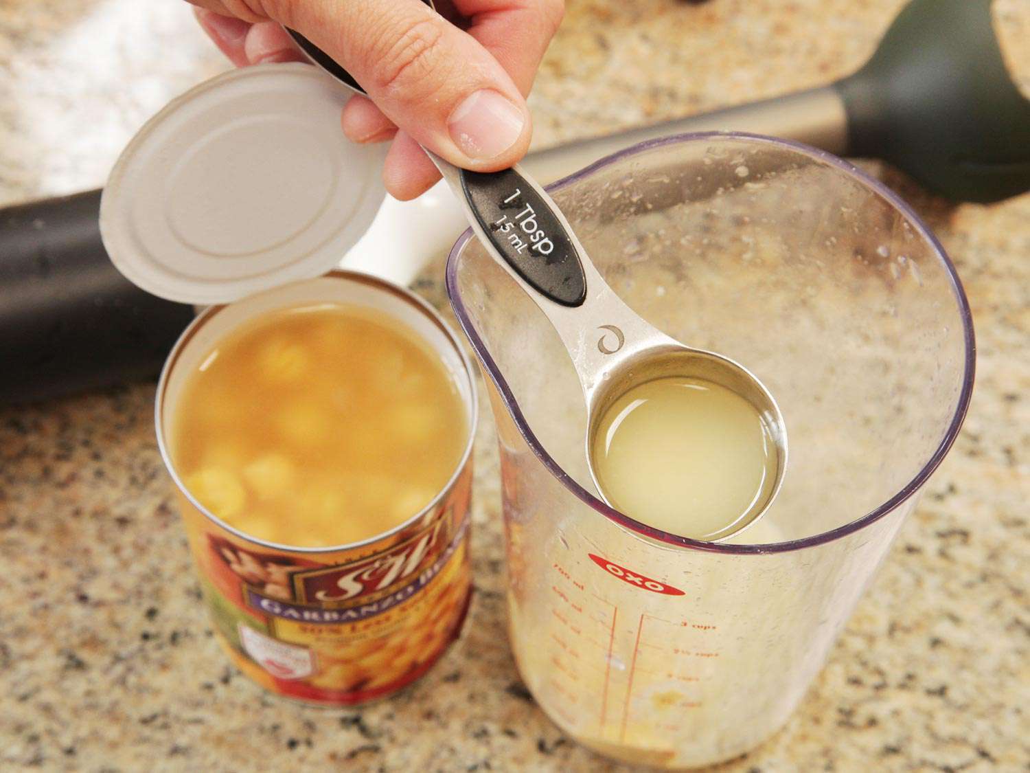 Adding a tablespoon of chickpea liquid to a jar, prepared to be blended with an immersion blender. 
