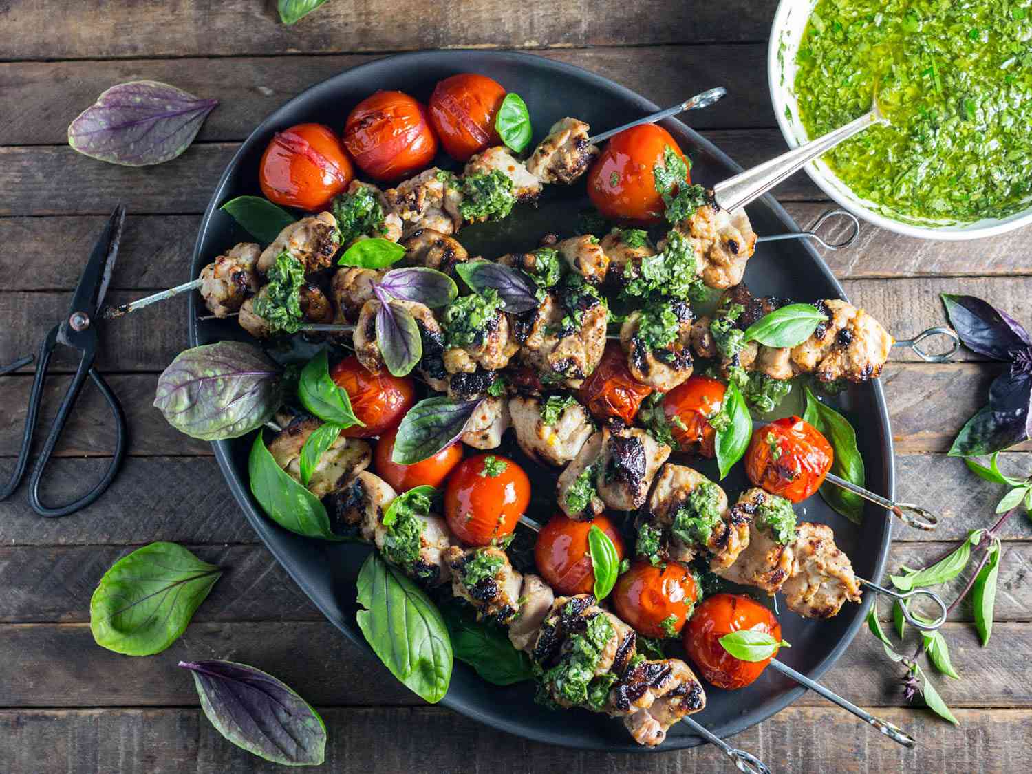 Overhead view of Grilled Lemon-Garlic Chicken and Tomato Kebabs With Basil Chimichurri, served on a black oval platter with extra chimichurri in a bowl nearby.