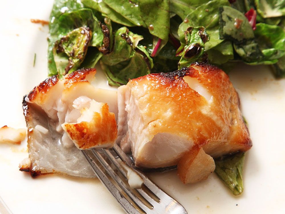 A half-eaten black cod fillet on a serving plate next to a pile of dressed greens. A bite of the fish is impaled on the tines of a fork.