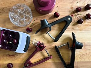 Five cherry pitters on a wooden surface with a few cherries strewn around them