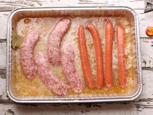 Overhead view of sausages and hot dogs partially submerged in a disposable aluminum pan filled with a flavorful braising liquid.