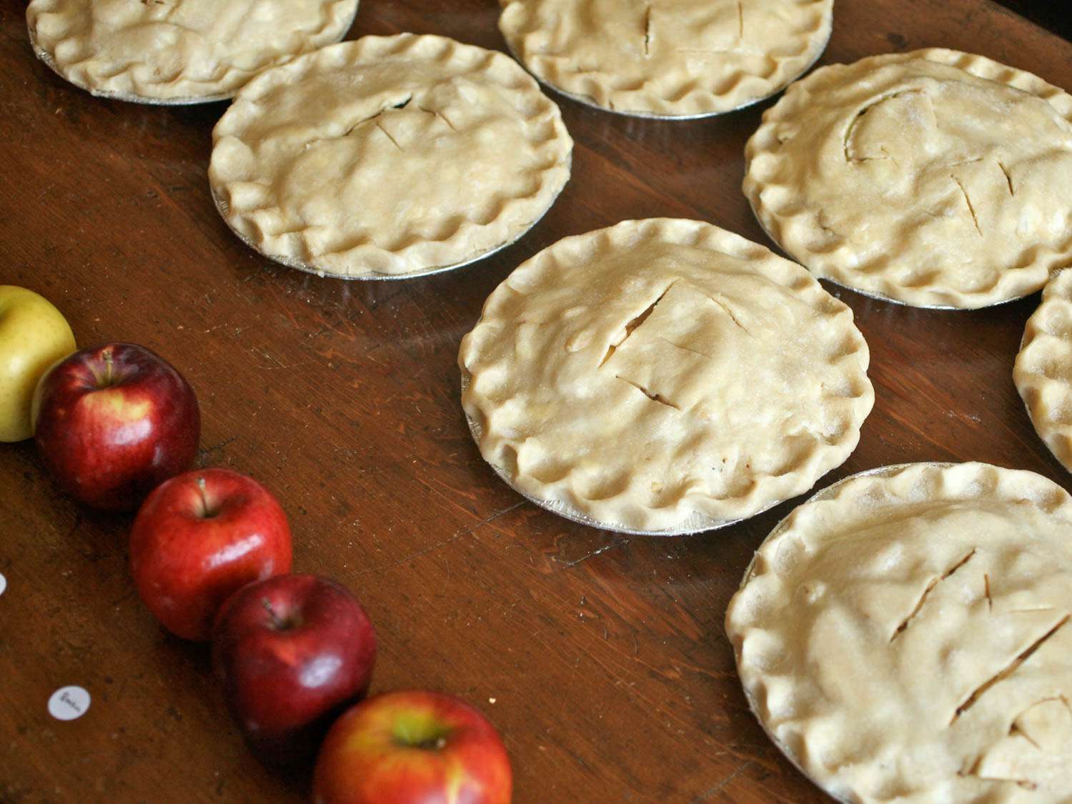 Several unbaked apple pies lined up on a wooden surface next to a row of apples