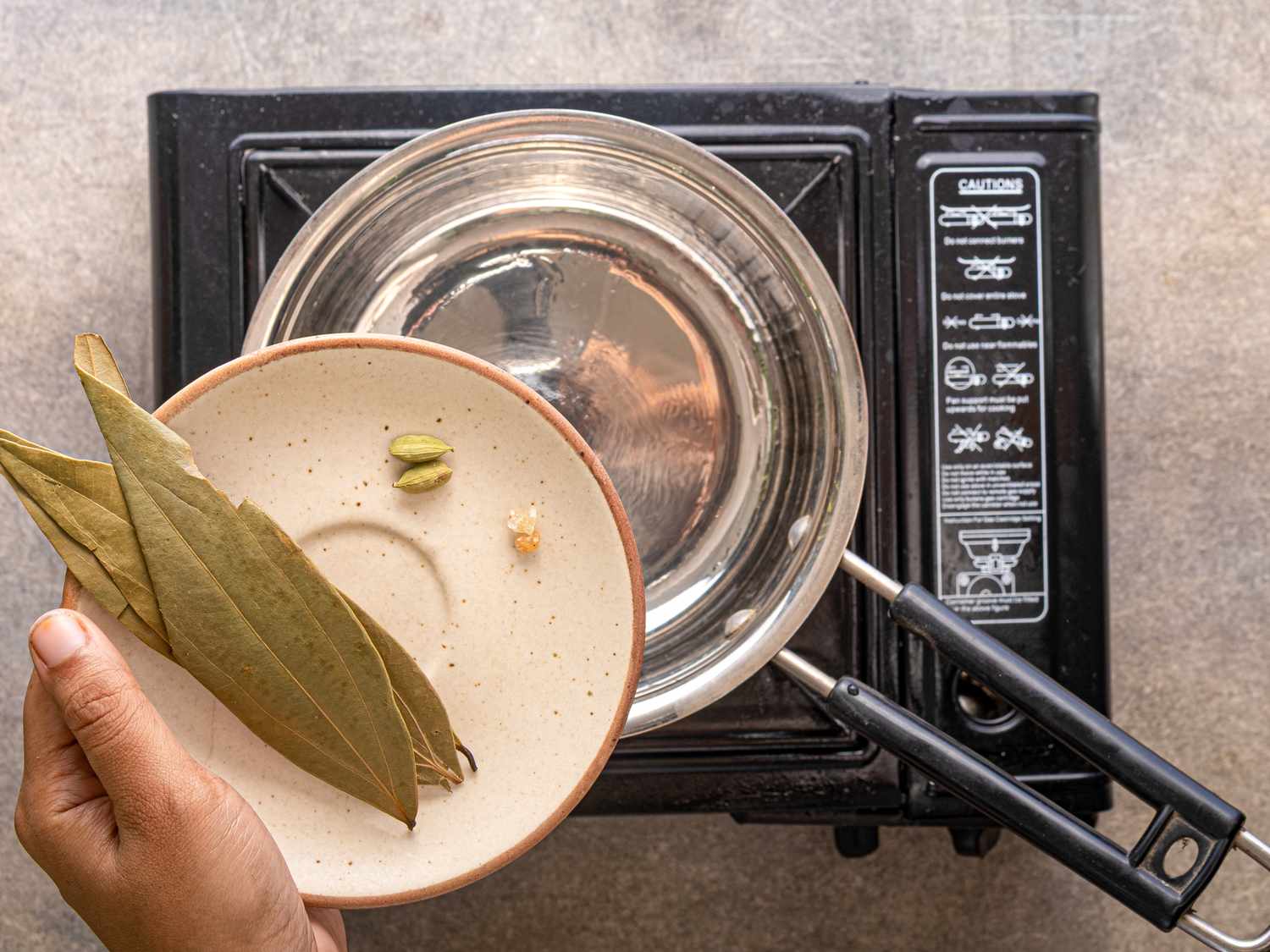 Hand holding a plate with bay leaves cardamom pods and a small piece of ginger above a cooking pot on a stovetop