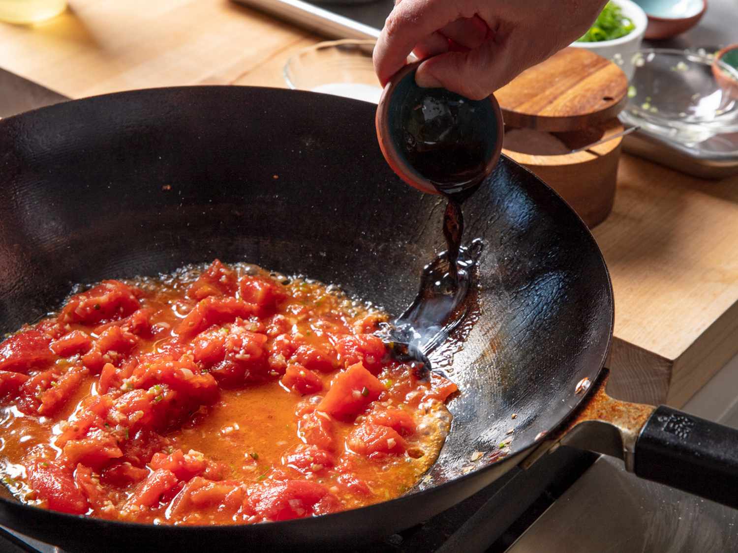 Soy sauce being poured near the top edge of a wok filled with tomatoes.