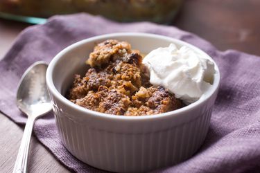 A white ramekin filled with apple crisp and whipped cream, next to spoon on a lavender cloth napkin.