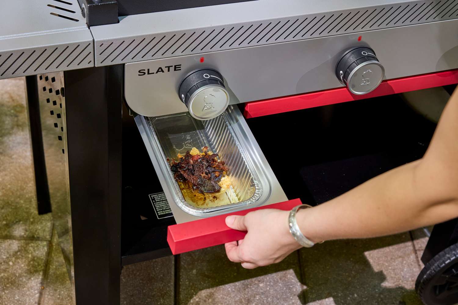 A person removes the grease trap from the Weber Slate 28 inch Rust-Resistant 3 Burner Outdoor Griddle