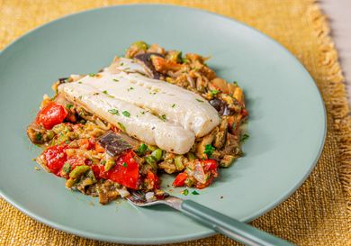 A plate of cod fillet served on a bed of caponata placed on a greencolored plate with a fork