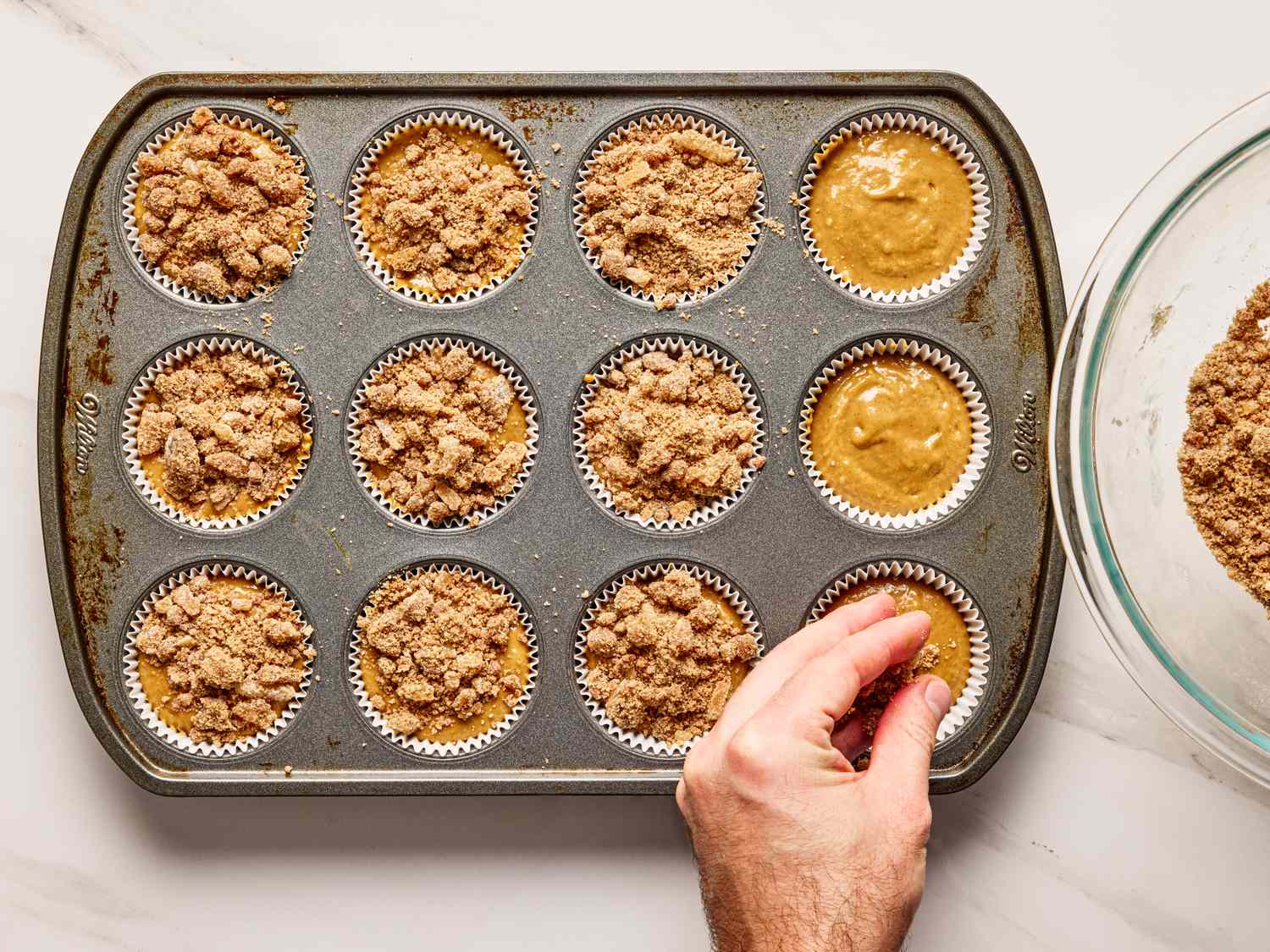 A hand placing crumb topping on gingerbread muffin batter in a muffin tin