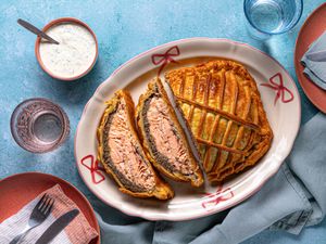 Salmon encased in pastry on a serving dish with dill sauce and tableware on a blue surface