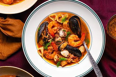 A seafood soup served in a white bowl with a spoon garnished with shrimp mussels and herbs placed on a maroon tablecloth