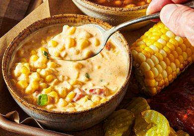 A bowl of cheesy corn garnished with diced vegetables served alongside a cob of corn pickle slices and barbecue items