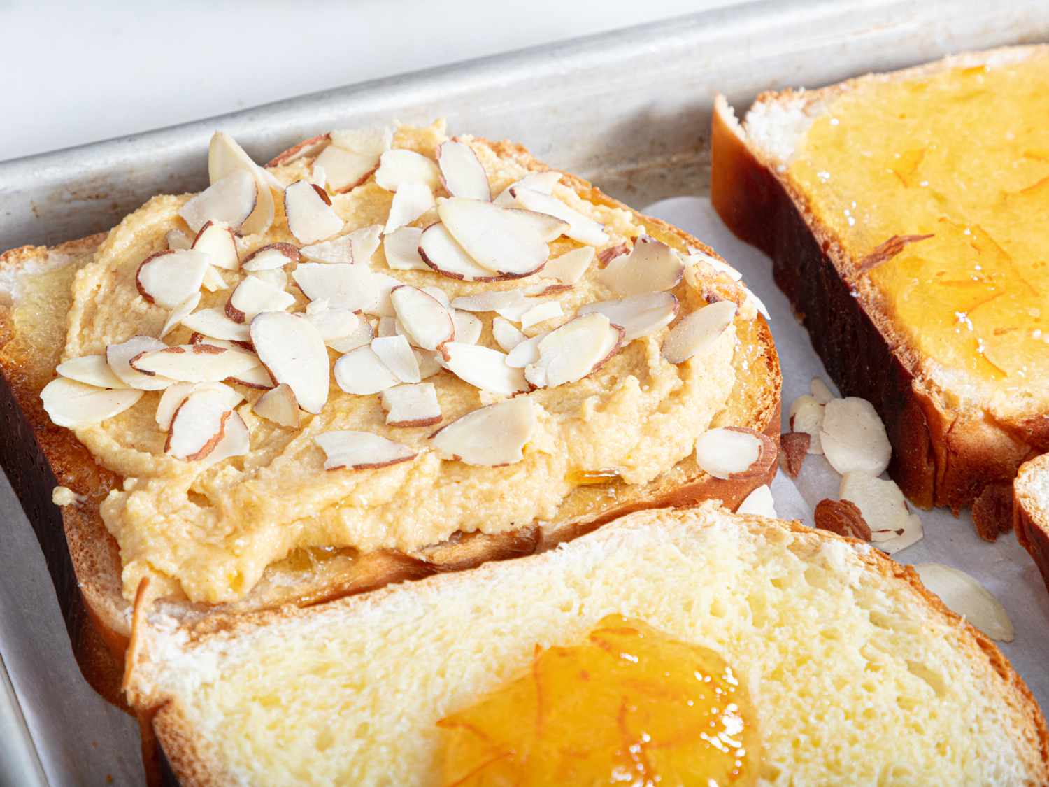3 slices of bread on a sheetpan. 2 slices have marmalade on them, and the 3rd had almond paste and almond slices on it 
