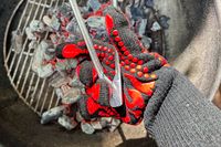 A person holds a pair of tongs over coals while wearing the Comsmart BBQ Gloves