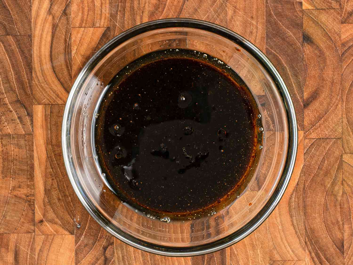  Sesame oil, light and dark soy sauces, oyster sauce, salt, sugar, wine, and water mixed inside a small glass bowl on a wooden cutting board.