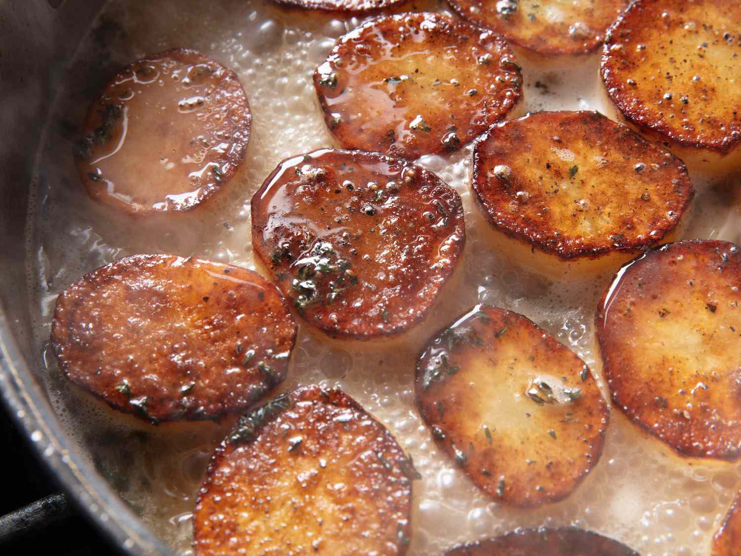 Closeup of potatoes simmering in a pan with stock, butter, thyme, and garlic