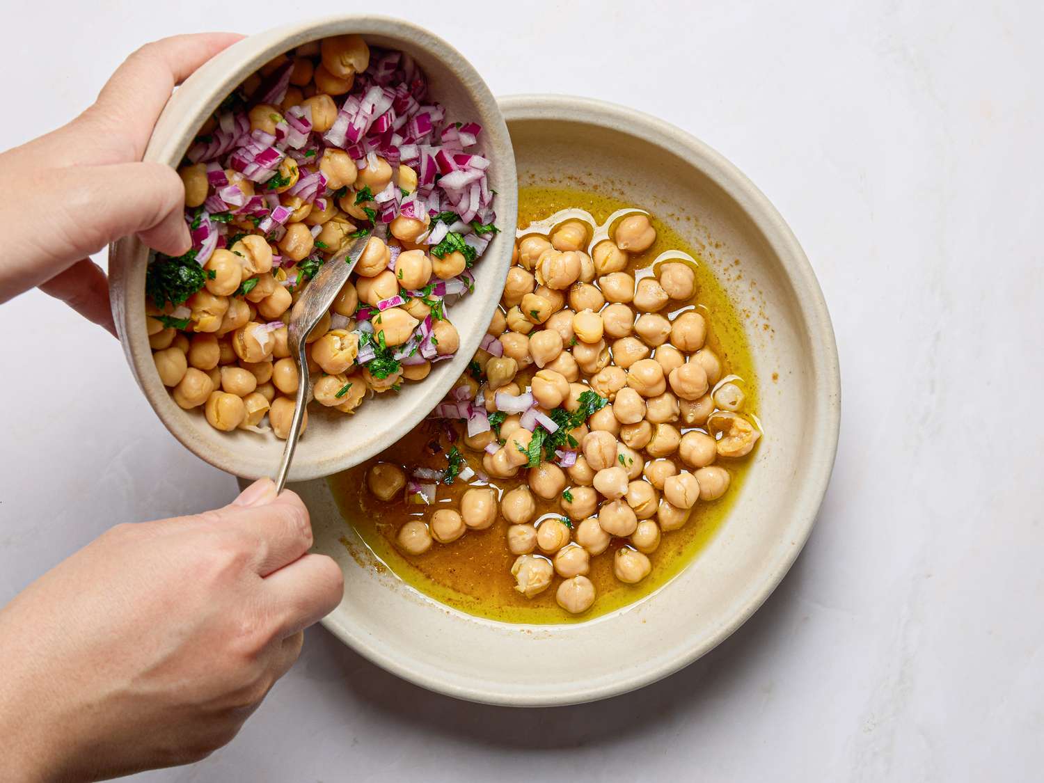 A bowl of chickpeas being poured into another dish with olive oil and herbs preparation of a chickpea salad