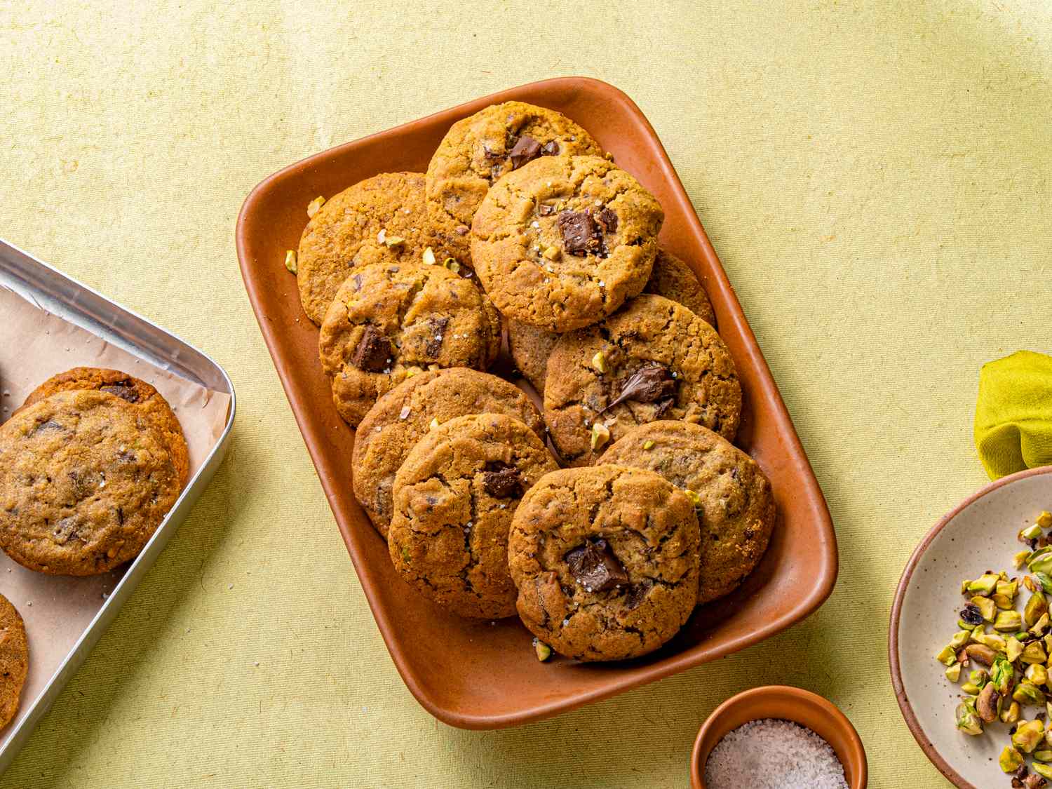 A tray of pistachio chocolate chip cookies with pistachios and salt in bowls nearby