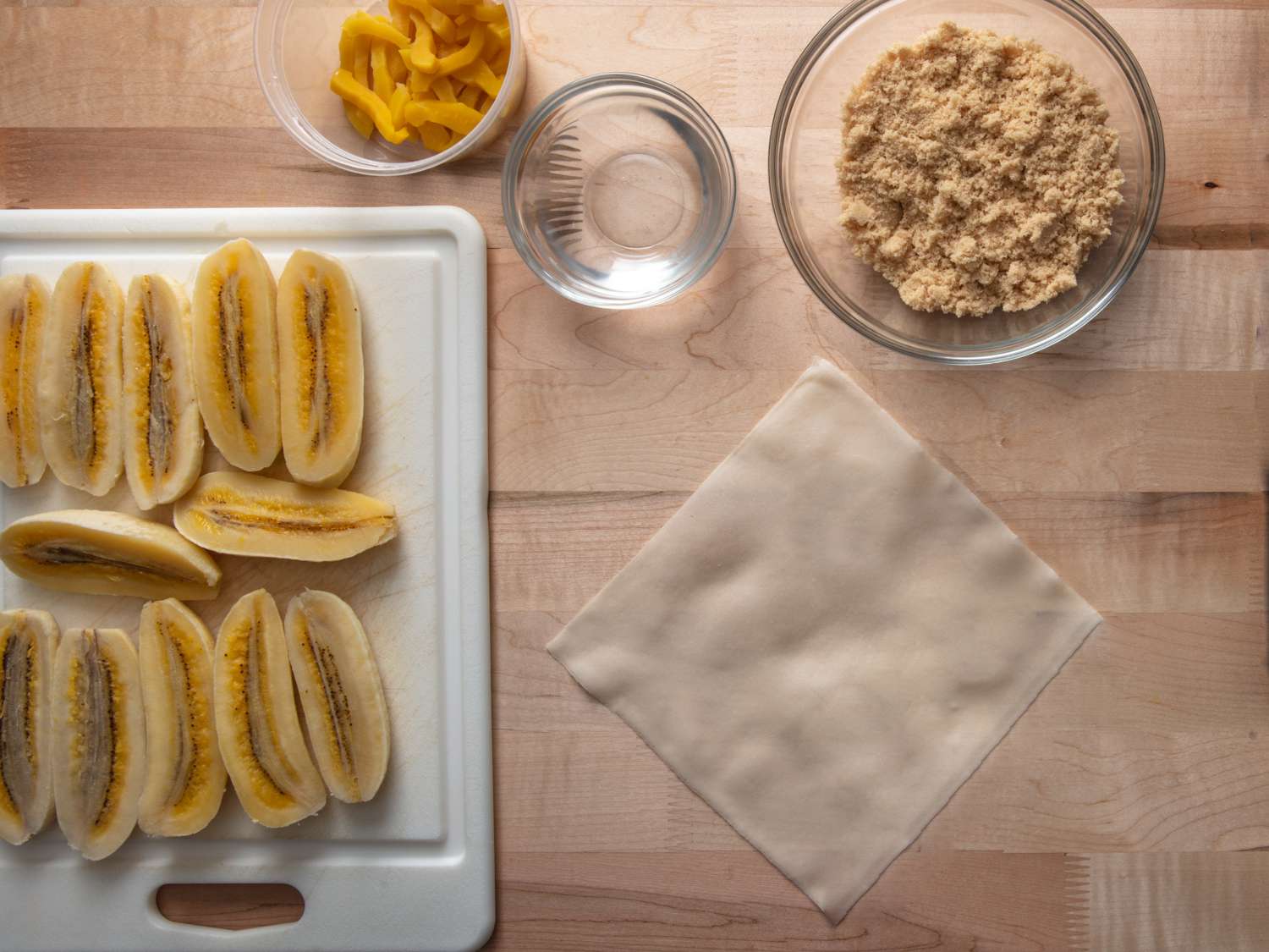 a workstation for making turon: saba bananas, brown sugar, jackfruit, a bowl of water, and a spring roll wrapper