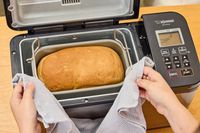 A person using a towel to remove a filled basket from the Zojirushi Home Bakery Virtuoso Plus Bread Maker