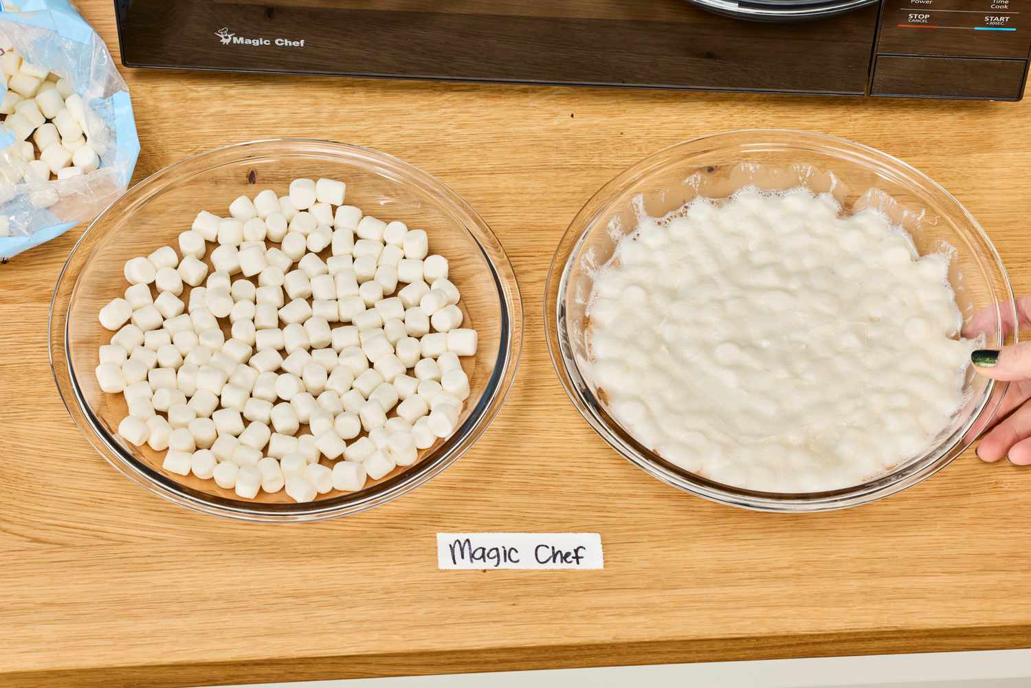 A hand placing a bowl of Magic Chef 1.1 Microwave heated marshmallows next to a bowl of unheated marshmallows on a wooden surface