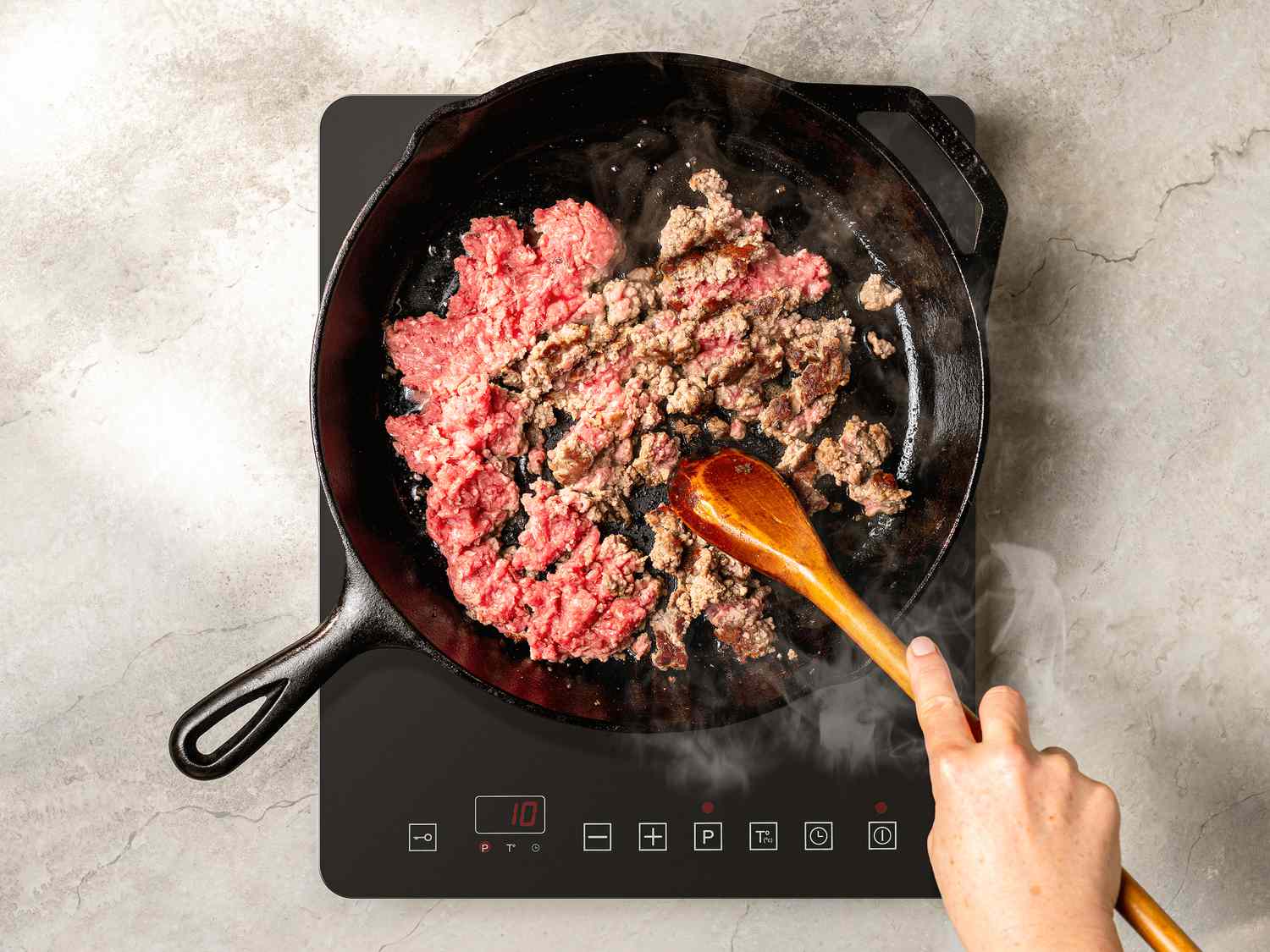 Ground beef cooking in a cast iron pan, being broken up with a wooden spoon