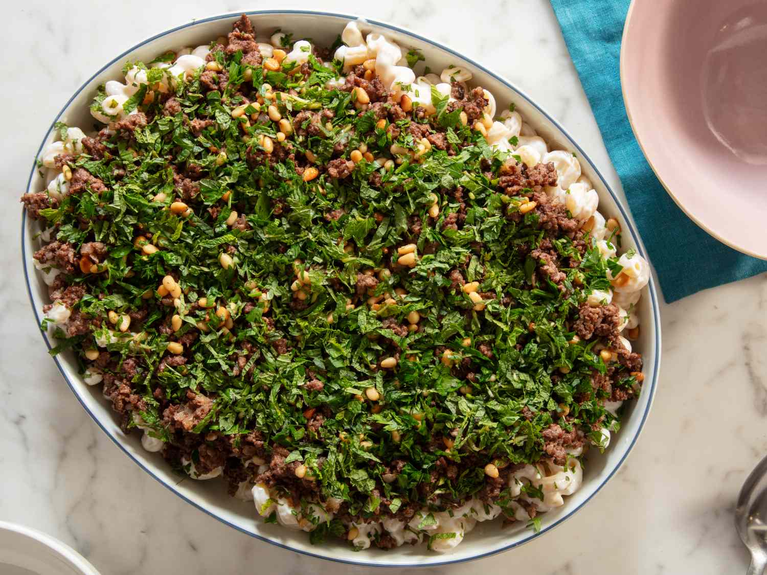 Oval dish filled with macaroni covered in cooked ground meat, herbs, and nuts, placed on a marble surface