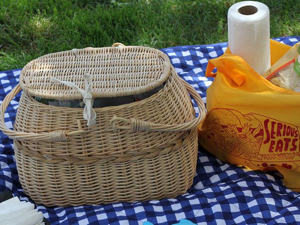 A wicker picnic basket on a blue checkered tablecloth spread on the grass outdoors. There is a Serious Eats bag with paper towels and other picnic supplies next to the basket. 