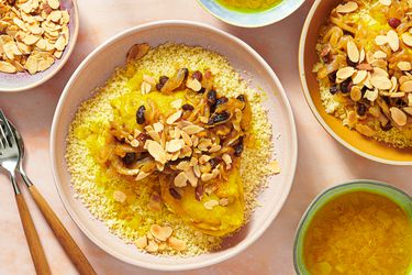 2 plates of Chicken couscous T'faya on pink surface surrounded by bowls of broth, toasted almonds, and forks