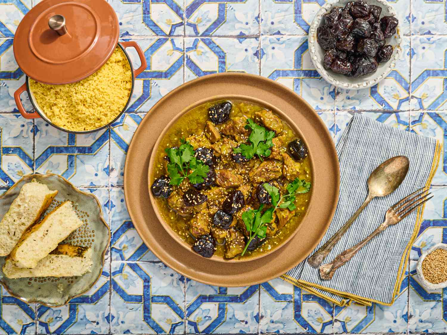 Lamb and prune Tagine with side of couscous, prunes, bread and cutlery on a blue tile surface 