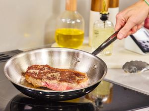 A hand holding a frying pan with a piece of steak being seared on a stovetop