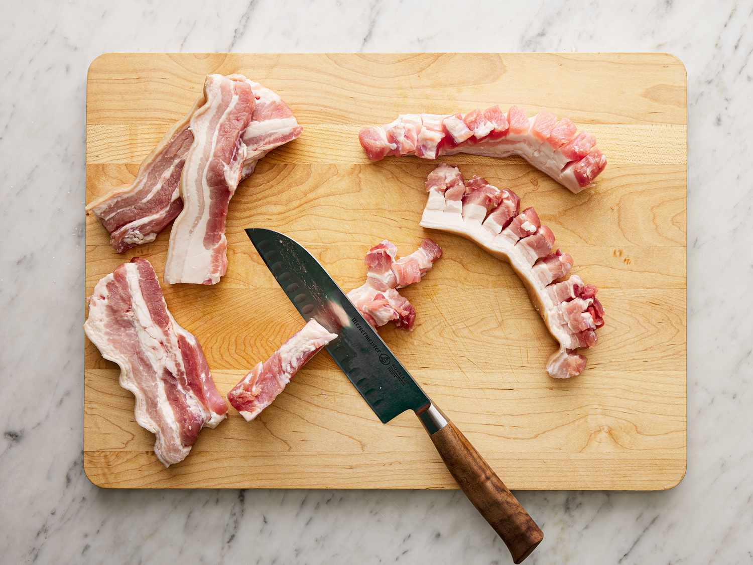 Overhead view of pork belly being cut with a chef's knife in half-inch intervals on a cutting board.