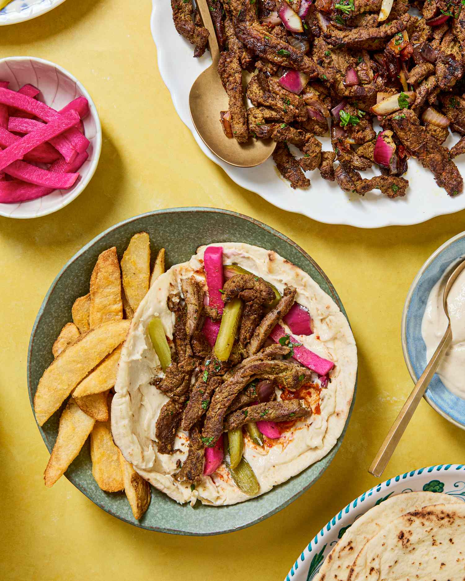 A plate with beef shawarma served with pickles and potato wedges surrounded by assorted bowls of Arabic cuisine