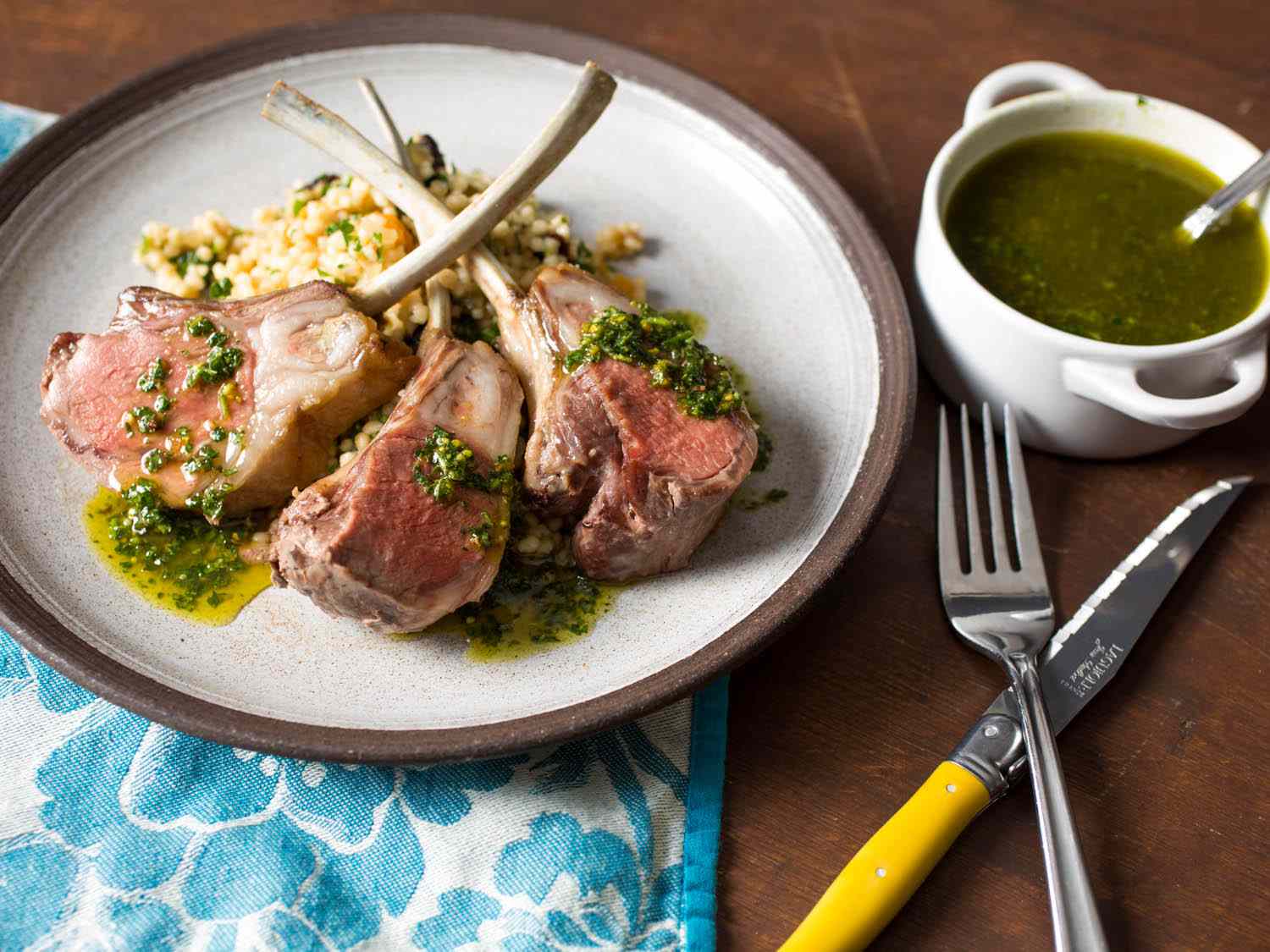 Lamb chops from a crown roast of lamb, on a plate with couscous stuffing, next to a small white crock of pistachio-mint sauce.