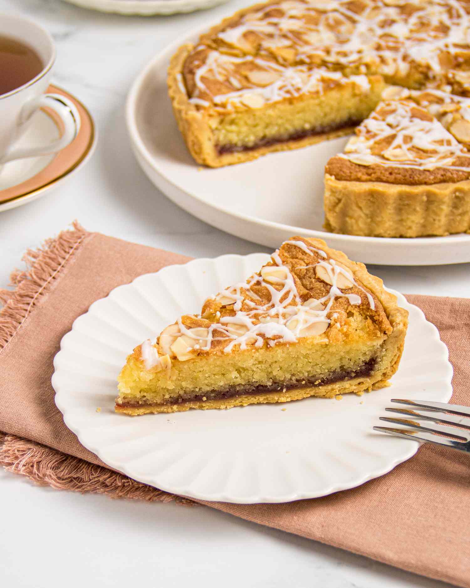 A slice of Bakewell Tart on a white plate, a larger tart in the background on a white table