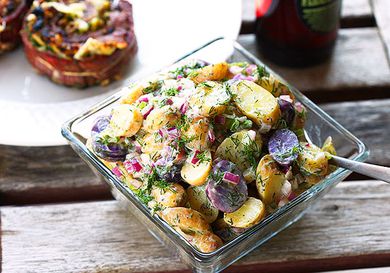 A square glass serving bowl filled with fingerling potato salad with creamy dill dressing is set next to grilled flank steak pinwheels on a weathered wooden table.